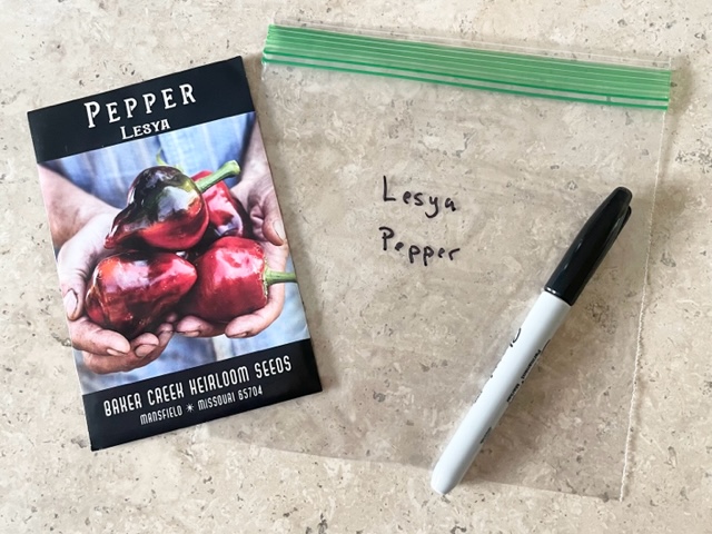 Packet of Lesya pepper seeds, a plastic sandwich bag labeled Lesya Pepper, and a parmanent marker on a marble table.
